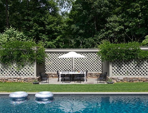 A beautiful gray-painted trellis style fence with climbing greenery offers privacy in this yard with a pool and outdoor patio with dining furniture and a white umbrella.