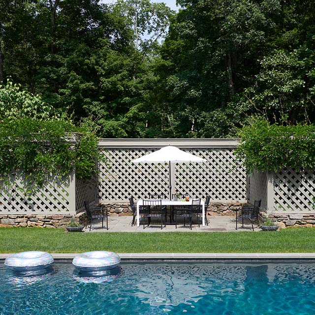 Une magnifique clôture grise de style treillage avec végétation grimpante préserve l’intimité de cette cour mettant en vedette une piscine et un patio, du mobilier de salle à manger et un parasol blanc.