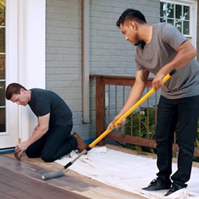 A gray brick home exterior with white trim and door has a deck being stained by two men: one man using a long roller, the other staining with a brush.
