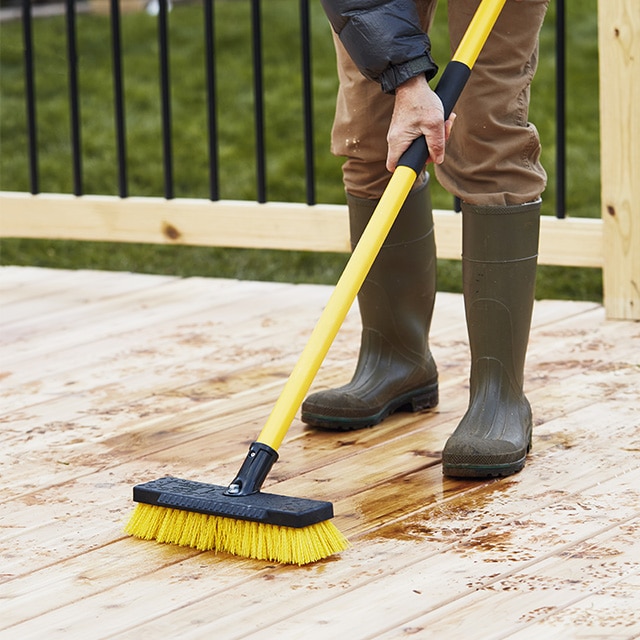 A man using a long yellow scrub brush on a new wood deck in preparation for staining and sealing.
