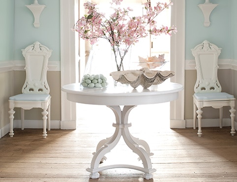 Una entrada con una mesa pintada de blanco con decoración floral, dos sillas blancas y paredes de color celeste, marcos blancos y revestimiento con paneles de madera beige.
