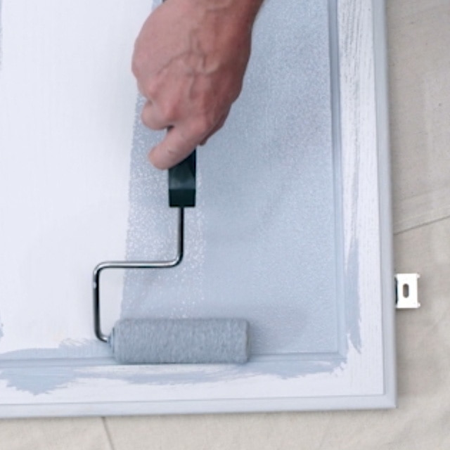 Una persona aplicando con rodillo imprimador en una puerta de gabinete de cocina pintada de blanco que está apoyada horizontalmente sobre una lona blanca.