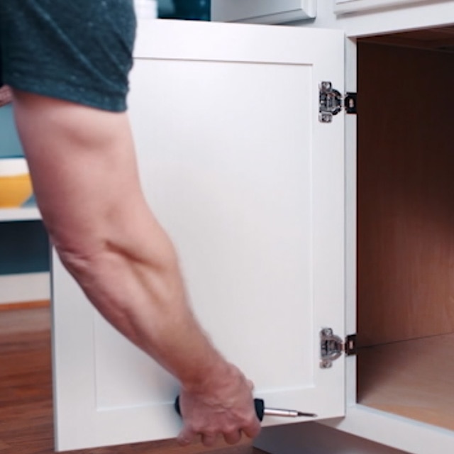 Una persona sosteniendo un destornillador y retirando una puerta de gabinete de cocina pintada de blanco, de sus bisagras.