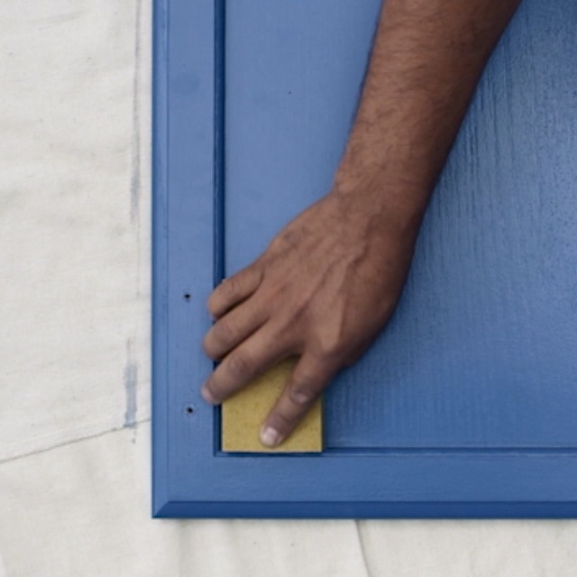 Una persona lijando una puerta de gabinete de cocina pintada de azul profundo sobre una lona blanca, antes de la segunda capa.