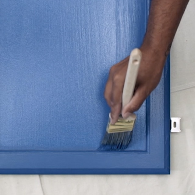 Una persona aplicando con brocha pintura de color azul profundo en una puerta de gabinete de cocina que está apoyada horizontalmente sobre una lona blanca.