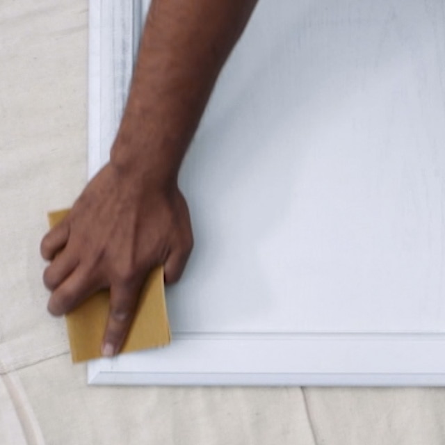 Una persona lijando una puerta de gabinete de cocina pintada de blanco que está apoyada horizontalmente sobre una lona blanca.