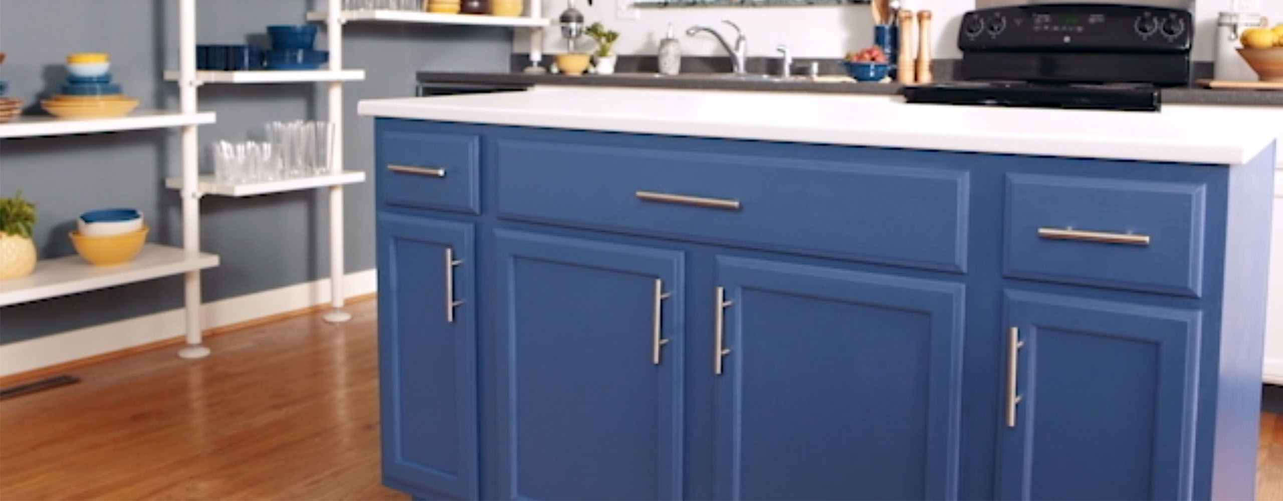 Esta cocina luminosa cuenta con una pared lateral pintada en gris con estanterías abiertas blancas, una isla con gabinetes pintados en azul profundo y una encimera blanca, y una pared del fondo blanca.