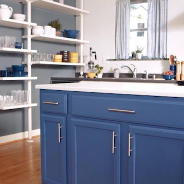 Esta cocina luminosa cuenta con una pared lateral pintada en gris con estanterías abiertas blancas, una isla con gabinetes pintados en azul profundo y una encimera blanca, y una pared del fondo blanca.