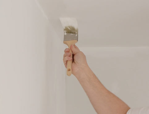 A closeup of a person painting a white ceiling corner with a paintbrush.