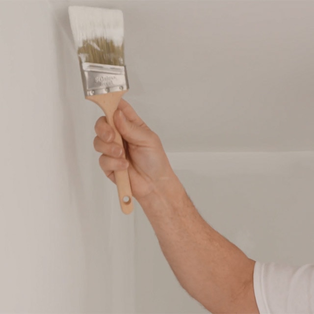 A closeup of a person painting the edge of a white ceiling with a paintbrush.