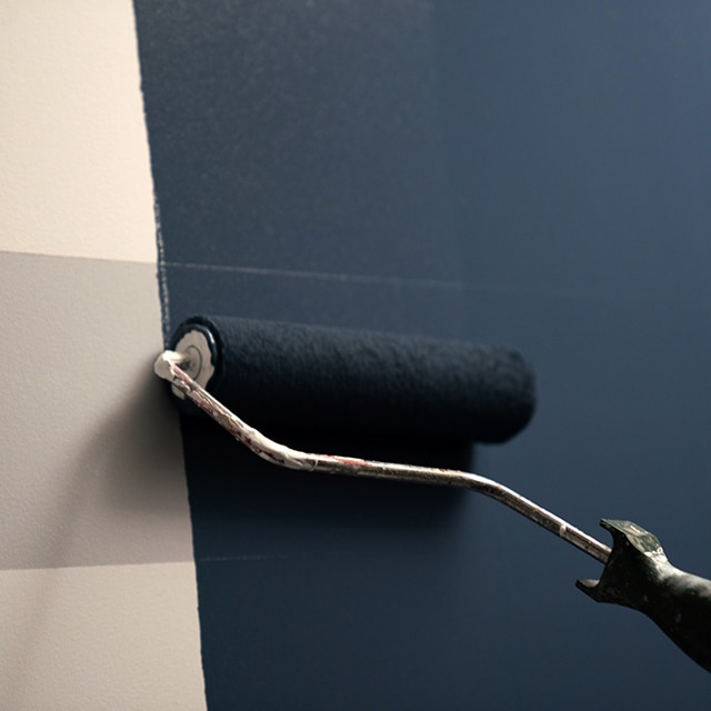 A white and gray striped wall being painted with AURA Interior paint in a dark blue using a roller.