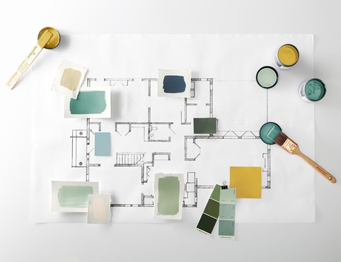 A bird’s eye view sees a variety of Benjamin Moore color swatches, color chips and 8 oz. color samples, plus a wooden paint mixer and a paint brush, atop a printed floor plan of a building.